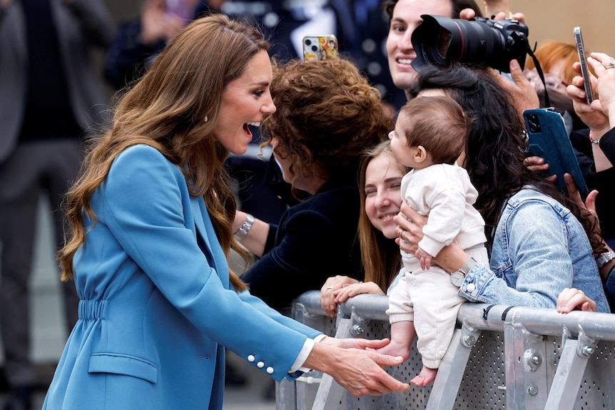 Princess Kate greeting a baby being held over a barrier by a mother in a crowd.