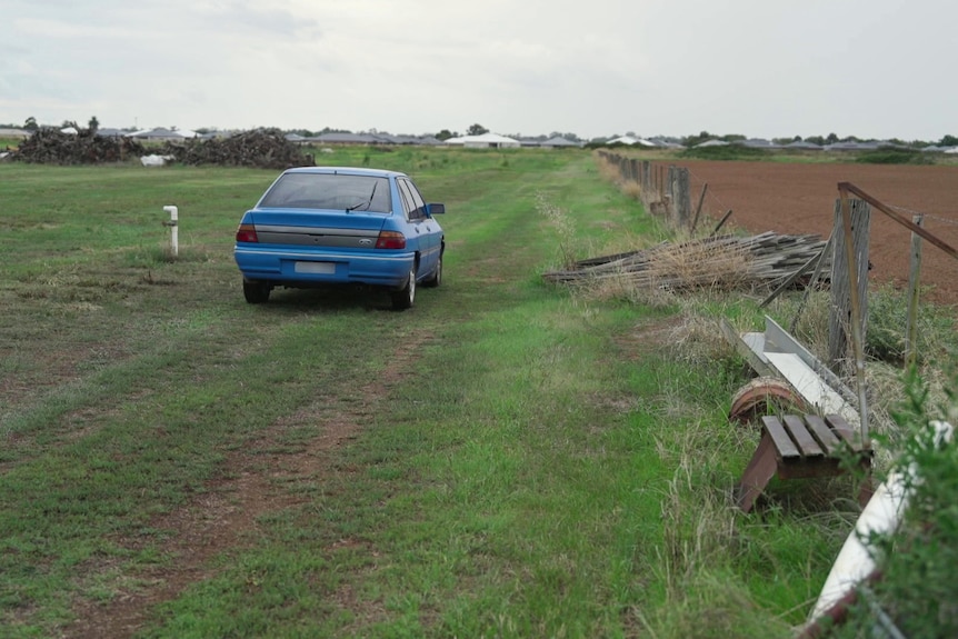 A blue 1990s Ford is parked on green grass in a paddock next to a fence that has fallen.