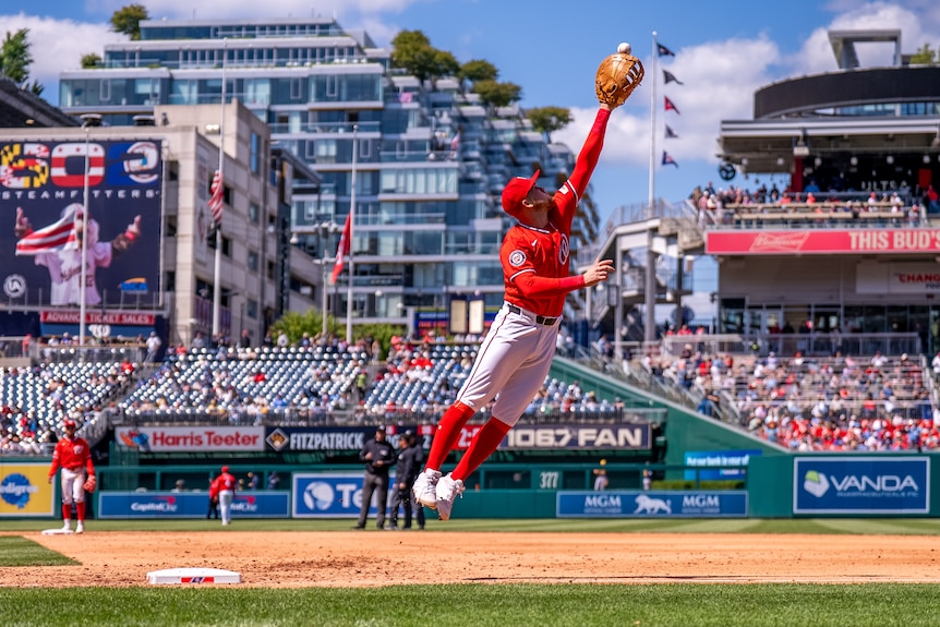 Curtis Mead, wearing red kit, jumps into the air to catch a ball in his baseball mitt on the field.