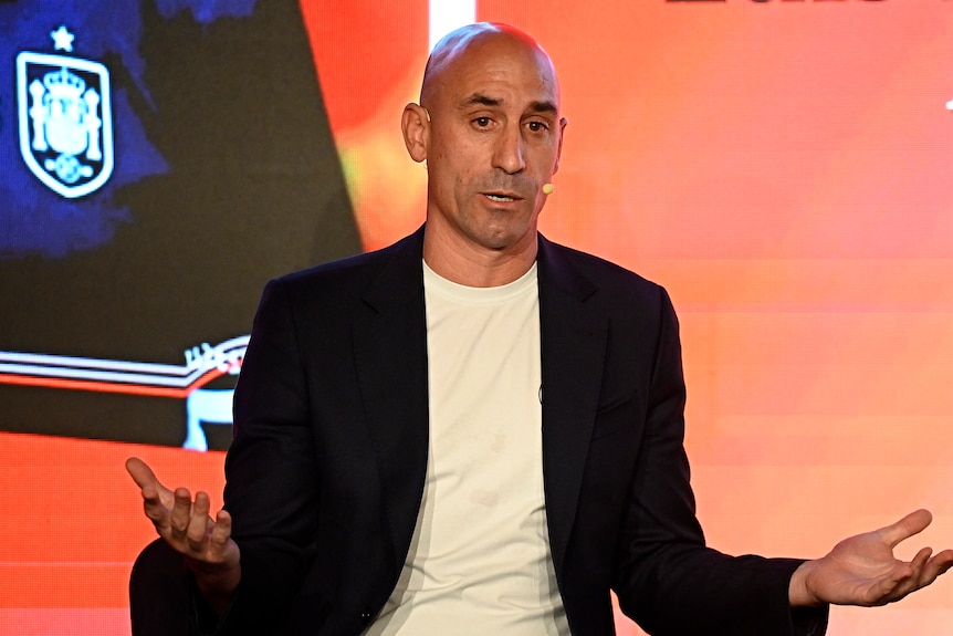 Luis Rubiales, wearing a T-shirt and sport coat, sits and speaks while gesturing with his hands.
