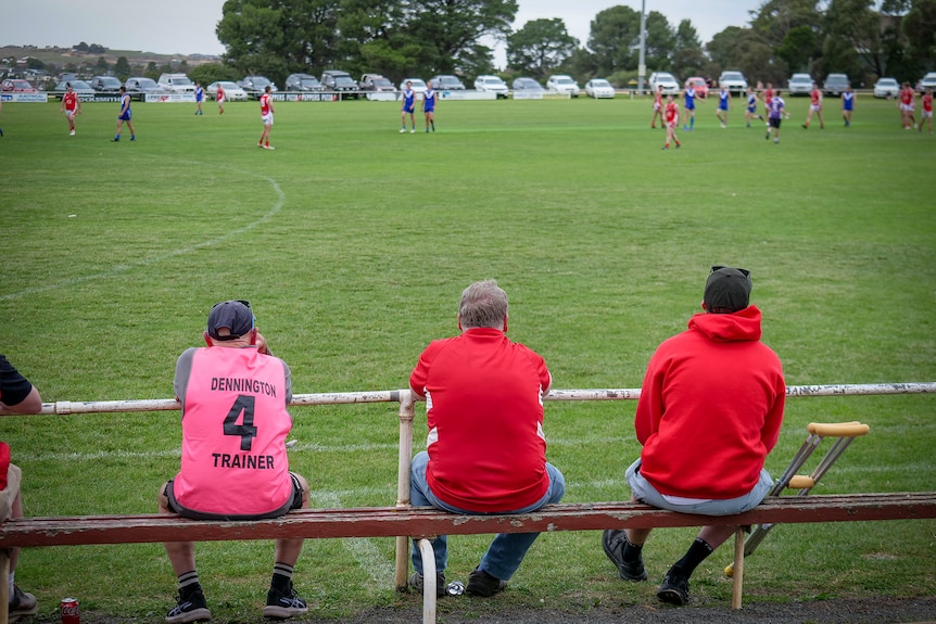 Three men in red watch a country football match. One has crutches next to him.