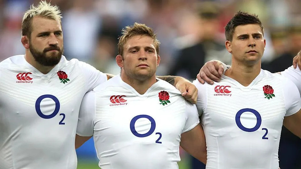 Three men in white England rugby kits stand with arms round shoulders and stare intently off camera. Left to right is a man with dyed blond spiky hair and a brown beard, a shorter man with blond hair and a slightly taller man with short brown hair.