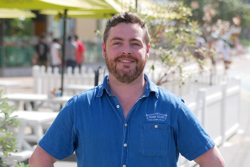 A man in a blue shirt smiles at the camera while standing outdoors near yellow umbrellas.