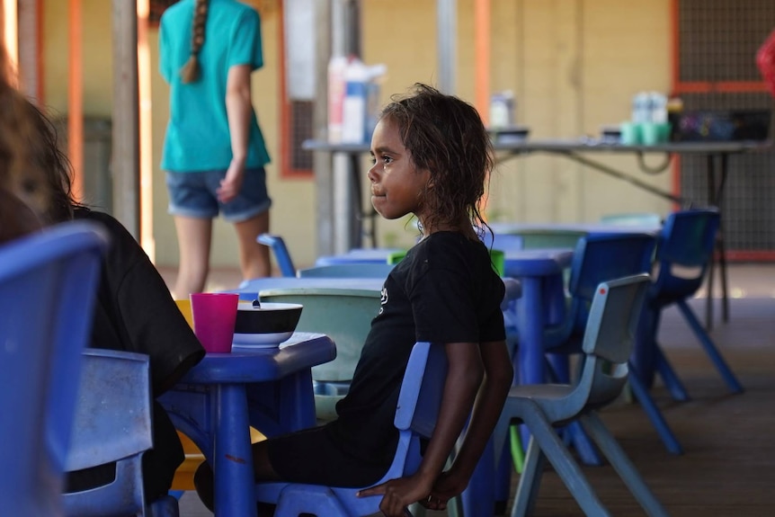 A young Aboriginal girl sits at a plastic table with a bowl and cup in front of her.