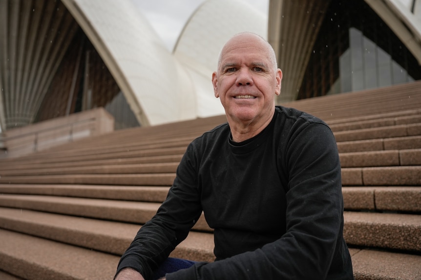 Michael, a bald Indigenous man sitting on the steps of the Sydney Opera House, smiling.