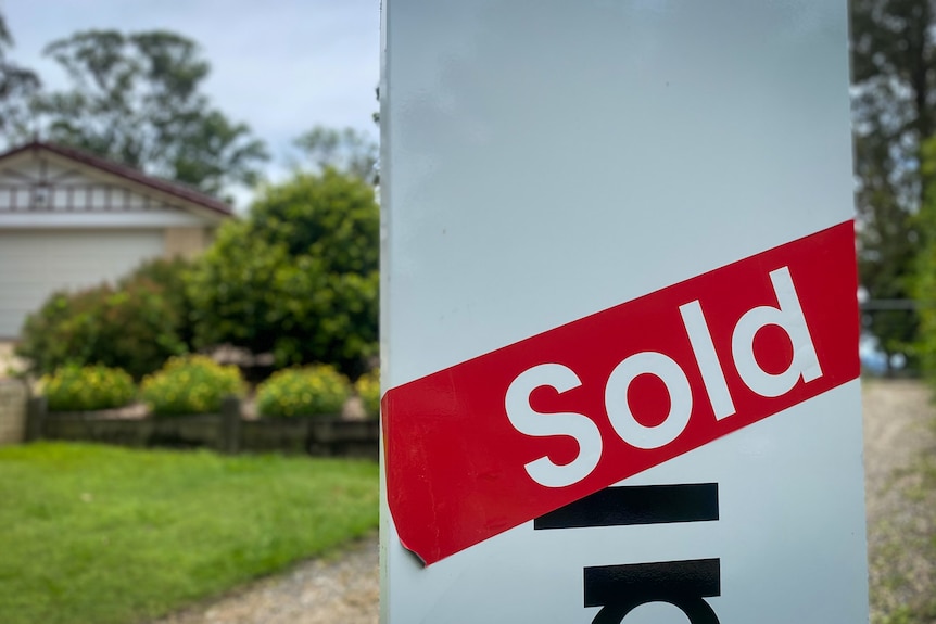 A red sold sticker across a for sale sign outside a house with a large front yard on a sunny day