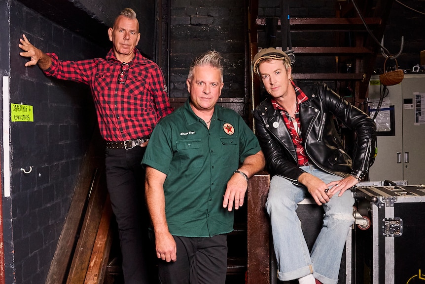 Three middle-aged men in rock 'n' roll outfits pose backstage in a theatre