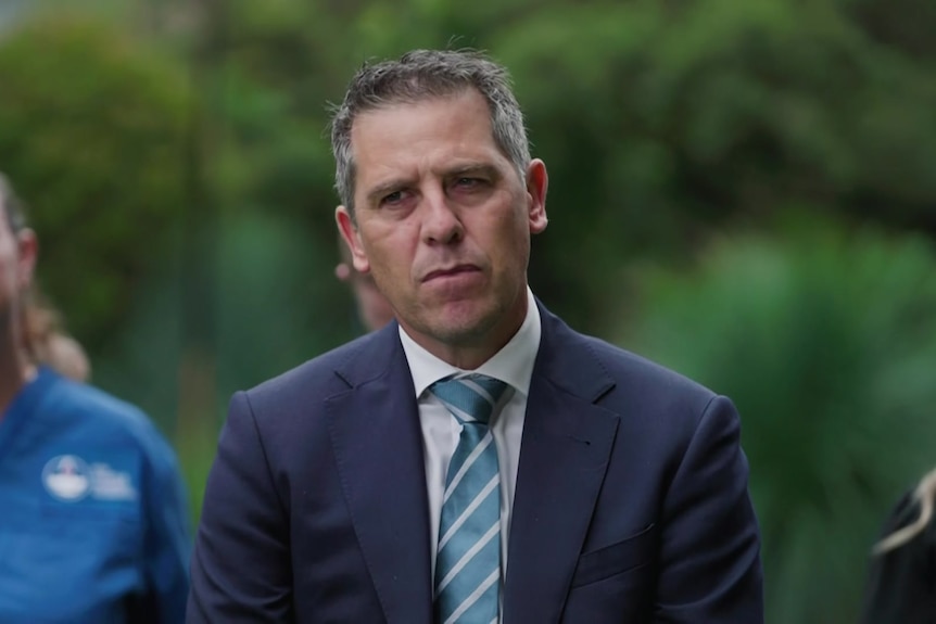 A man with grey hair in a blue jacket, white collared shirt and teal tie with white stripes stands in front of trees.
