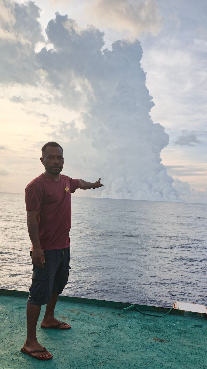 A man in a red shirt, shorts and flip flops stands on a boat pointing at a large plume of smoke in the disatance