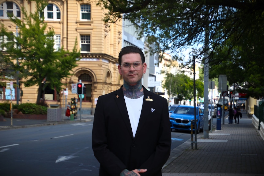 A heavily tattooed man wearing a white shirt and a black blazer stands on a busy street corner.