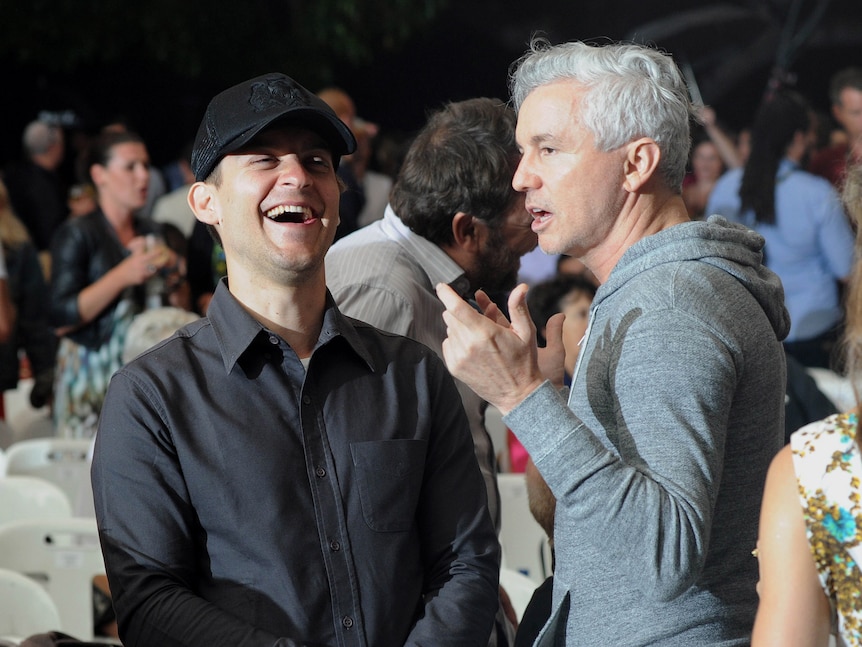 Tobey Maguire wearing a hat laughs big while talking to Baz Luhrmann, both standing at event