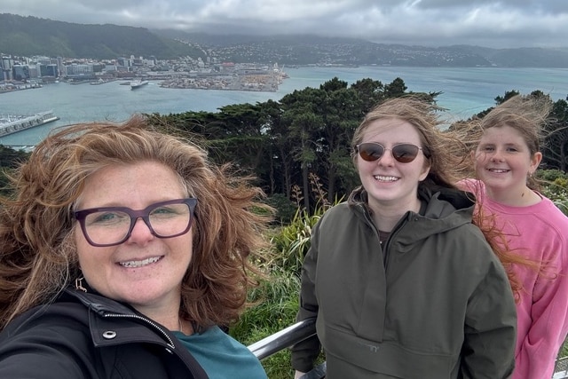 A woman takes a selfie with two smiling teenage girls on a hill overlooking a harbour.