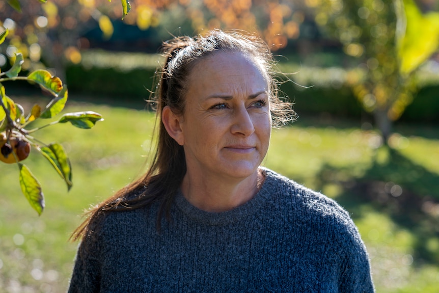 A woman in a pony tail and grey jumper is backlit in her front yard with orange and yellow leaves illuminated behind her.