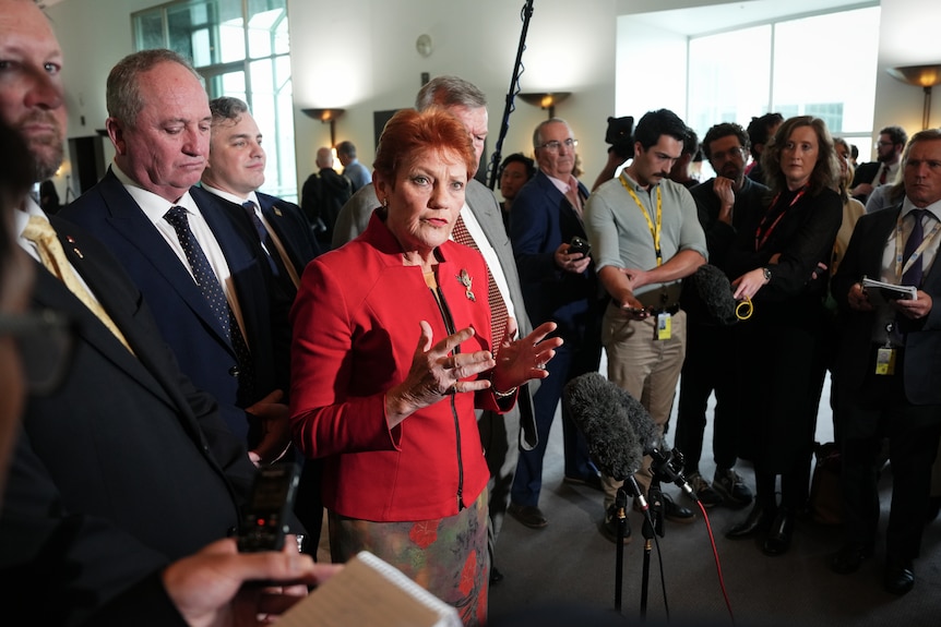 A woman in a red jacket, surrounded by people, speaks.