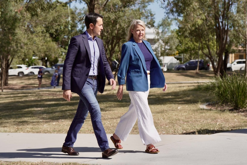 A blond woman in a blue suit and white pants outdoors and a man in a blue suit