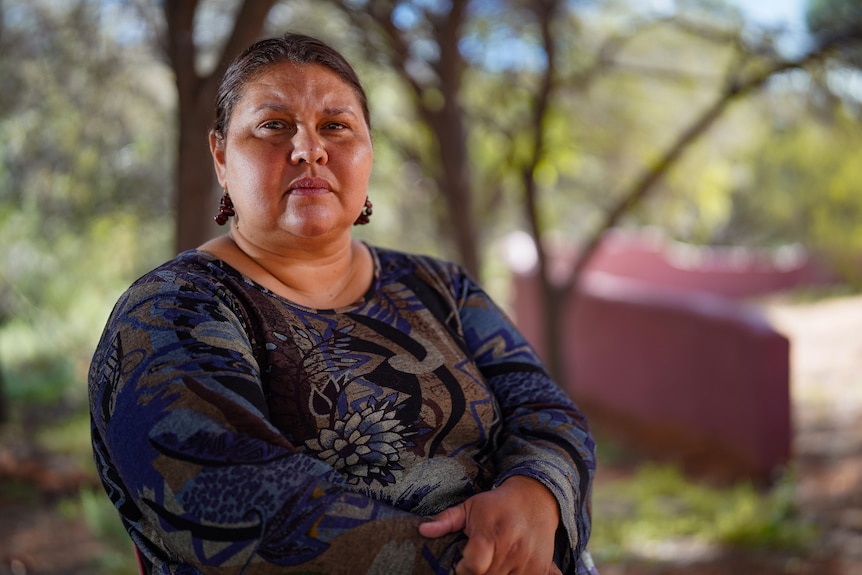 Aboriginal woman, brown/gray hair tied back, blue pattern deress, serious expression, sitting, blurred greenery behind her