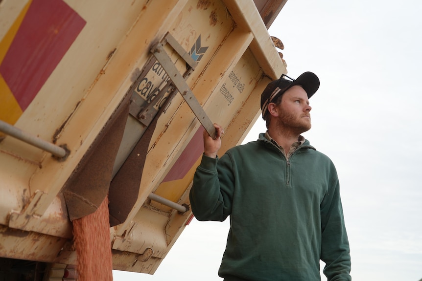 A man with a green jumper holding onto the back of a yellow truck.