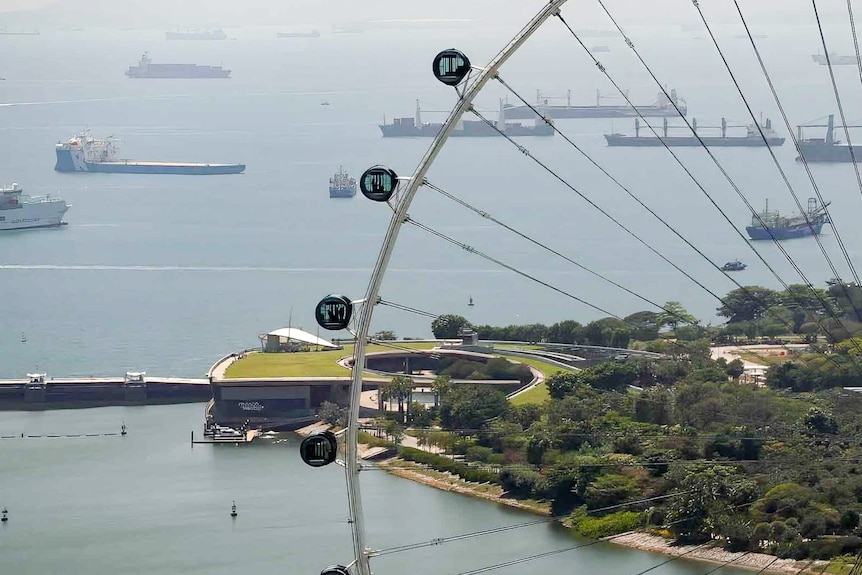 Cargo ships sitting just off the shore in Singapore, seen through the spokes of a ferris wheel.