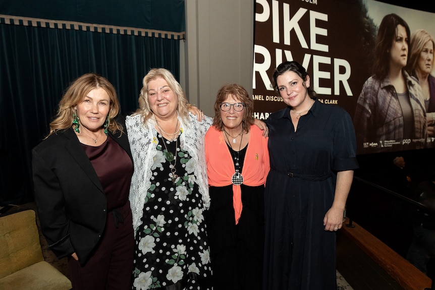 Four women stand in front of a large promotional poster saying Pike River