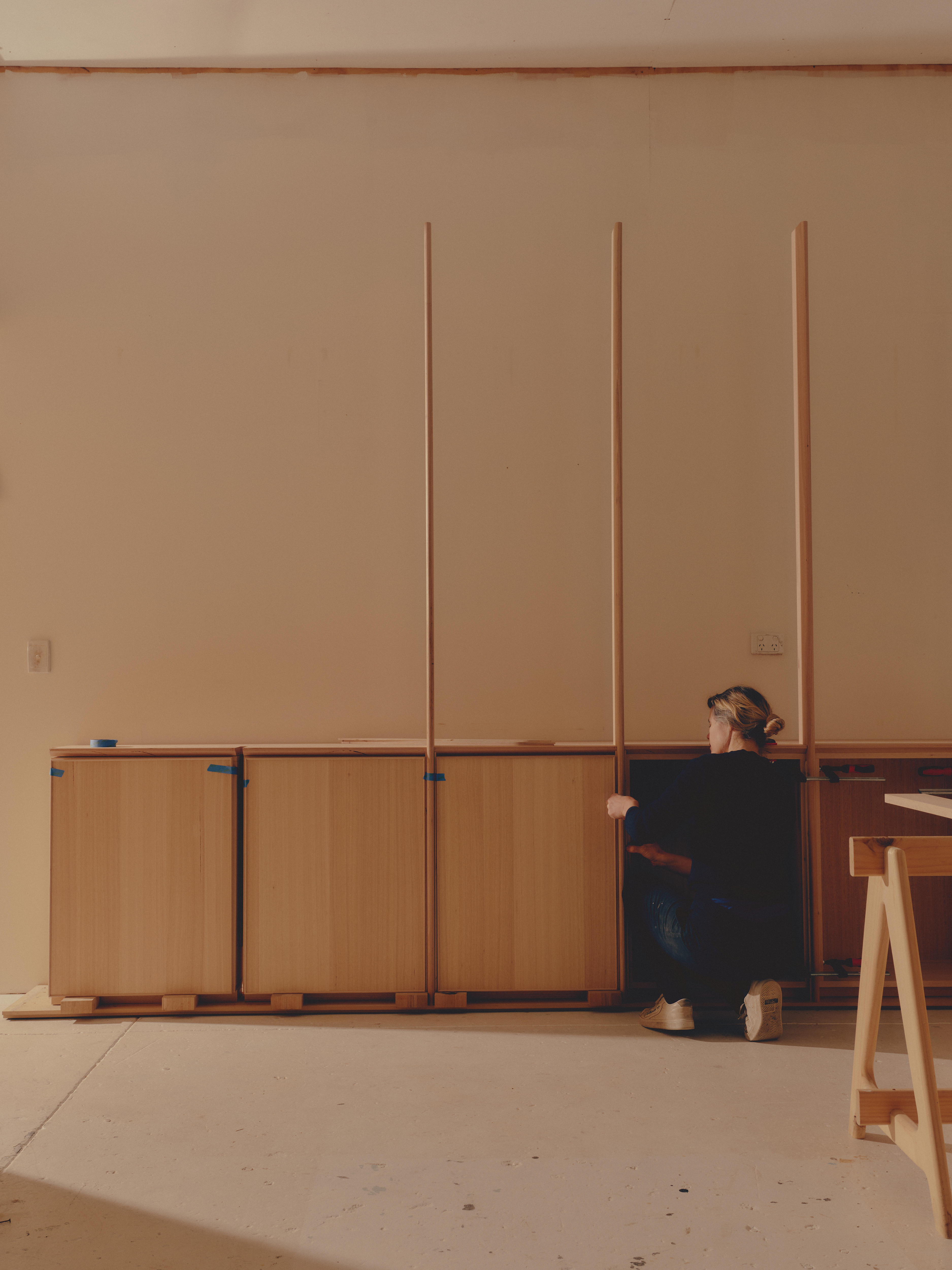 A woman crouches beside a partially made wooden cabinet.