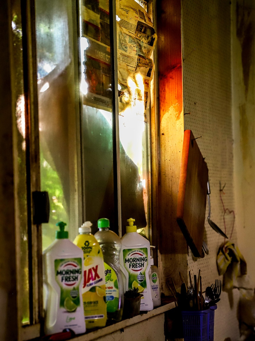 Light comes through a cracked window patched up with newspaper into a dirty kitchen with cleaning products in the foreground