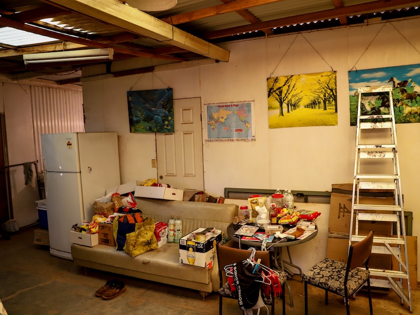 A dirty couch covered in various boxes and trash next to a ladder and fridge inside a derelict room