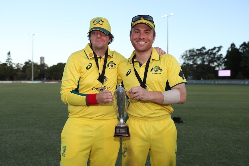 Two men in cricket clothing stand looking at the camera