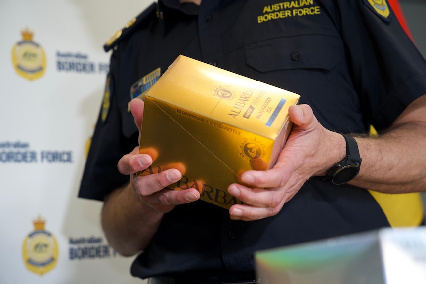 An Australian border force officer in uniform holds a box of vapes in gold packaging