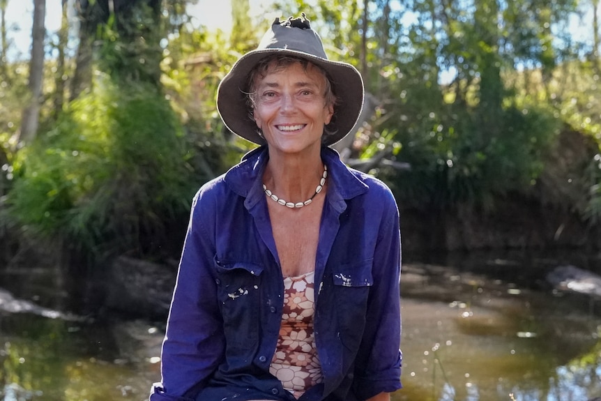 Rachel Ward wears a wide brim hat and long sleeve pull-over shirt while seated on a log in front of a brown creek and trees
