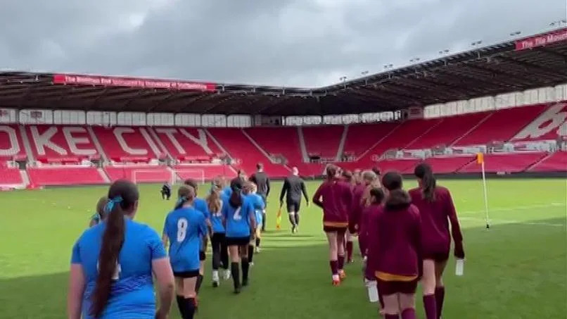 Two teams walking out onto the pitch in single file lines led by a referee. The pitch is surrounded by large red stands.