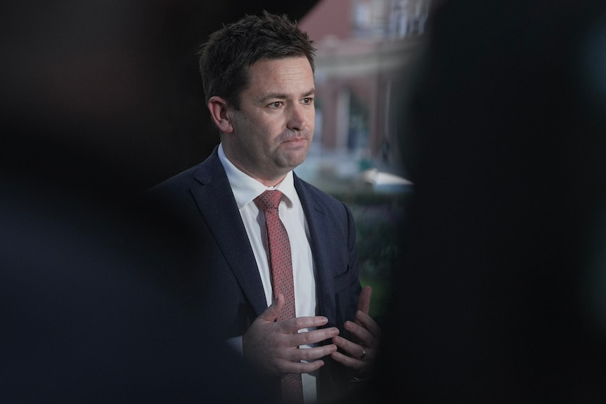 A man in a suit and tie gestures with his hands while speaking to media in a garden.