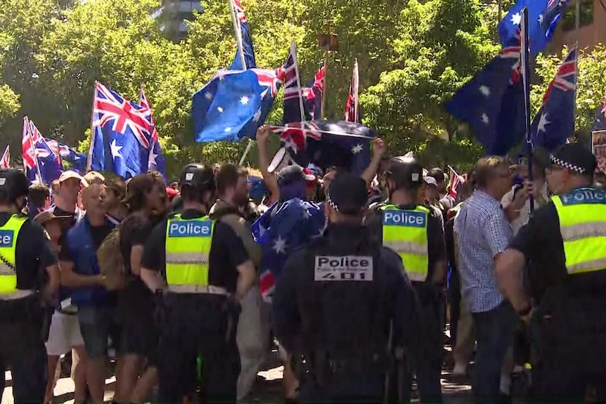 Police officers near a crowd of protesters waving Australian flags in a park.