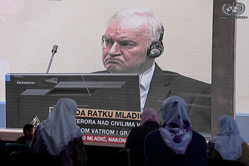 Several women wearing headscarfs watch a large TV screen, on which an older man wearing headphones sits behind a computer.