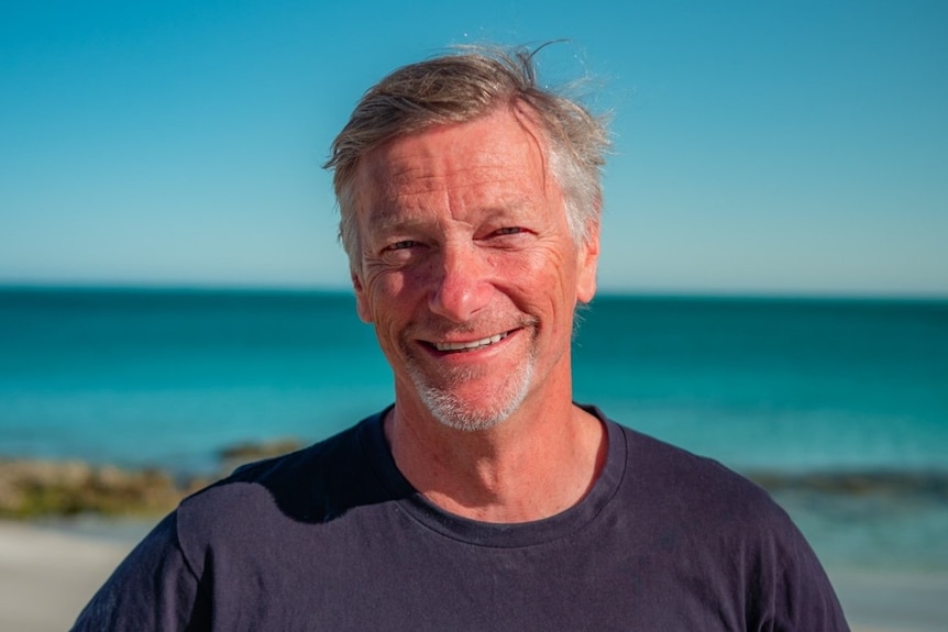 A smiling, middle aged man with grey hair stands in front of the ocean.