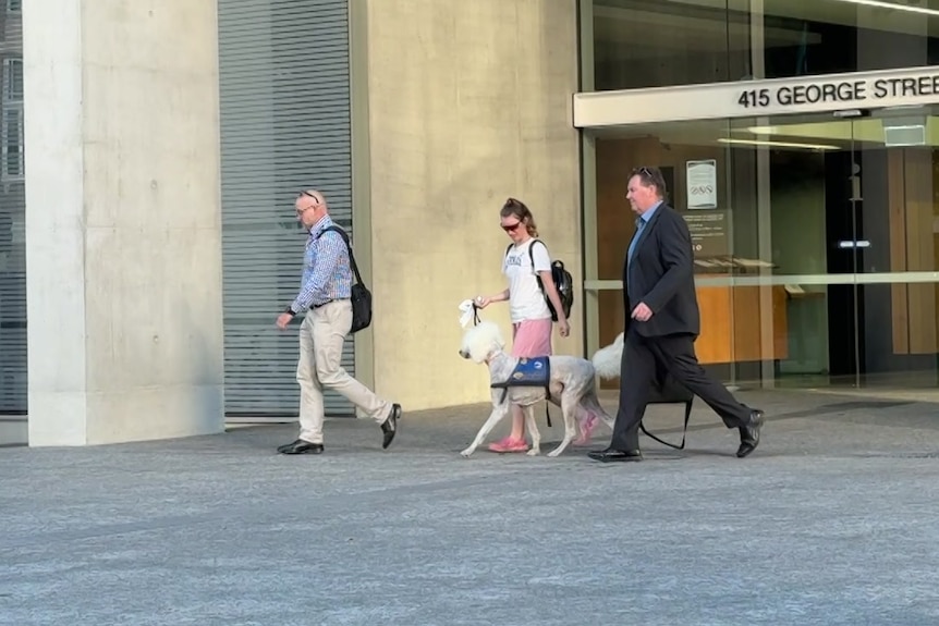 A woman with a dog walks out of a court building with two men either side of her.