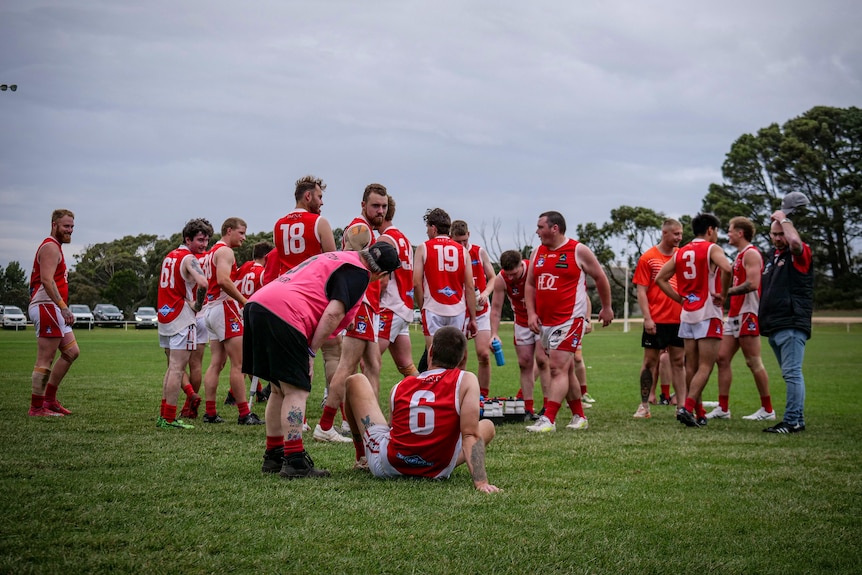 Footballers stand on the ground under grey skies. One man is sitting in the centre of the picture with his back to the camera.
