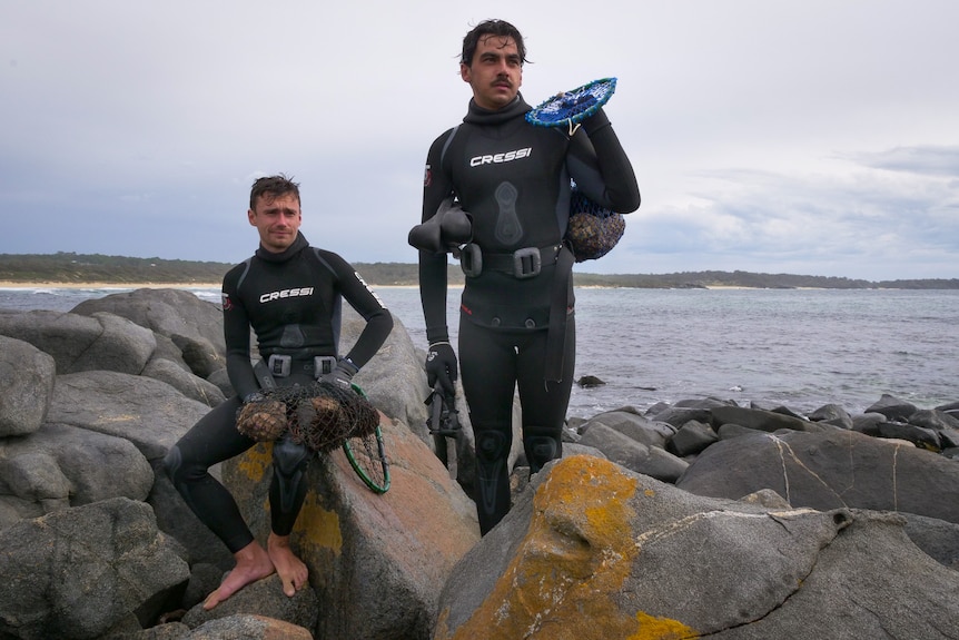 Two Aboriginal men stand in diving gear on rocks by the sea, holding their catch in net bags