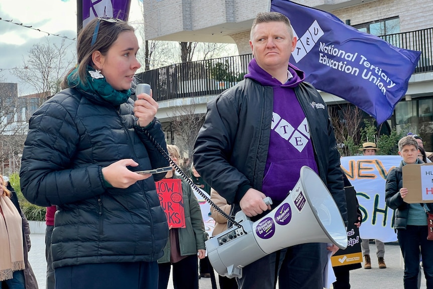 Two people stand next to each other looking serious, one holding a megaphone and the other speaking into its microphone.