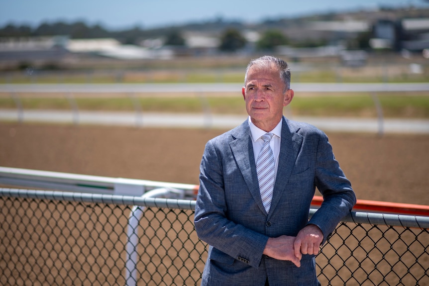 An older gentleman in a navy suit and striped white tie leans on the fence at the edge of a brown race track.