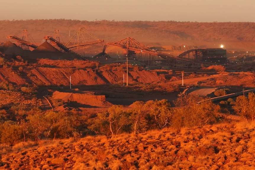 A view of a large mine site in the distance, several large pieces of equipment and mounds of iron ore can be seen.
