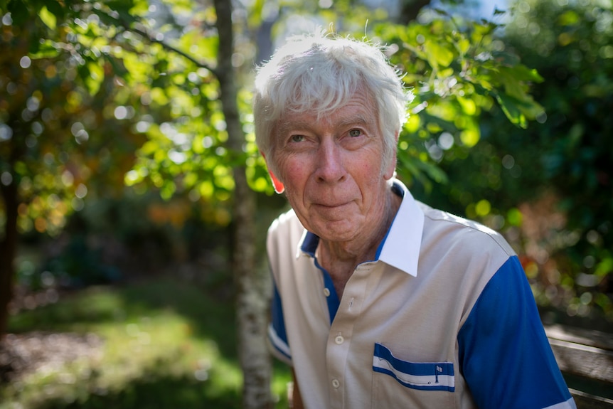 An elderly gentleman smiles in dappled light in a green leafy garden.