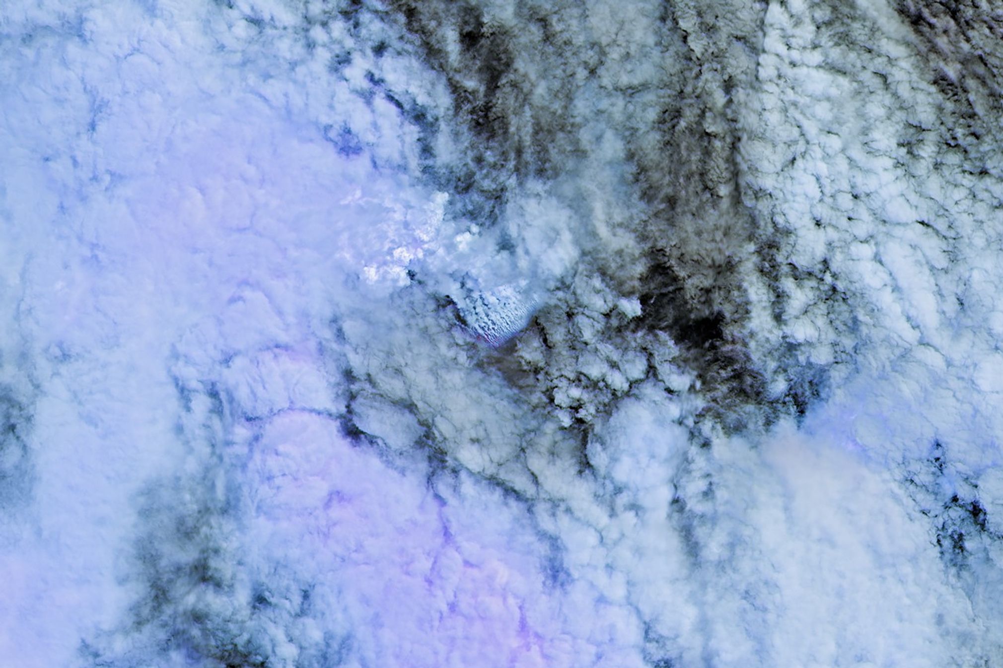 Only small patches of the ocean are visible through puffy, white volcanic plumes streaming from the center of the scene. Clouds not directly related to the eruption are visible throughout much of the image.  A false-color inset box shows the infrared signature of the eruption as a series of red dots near the volcanic plumes.