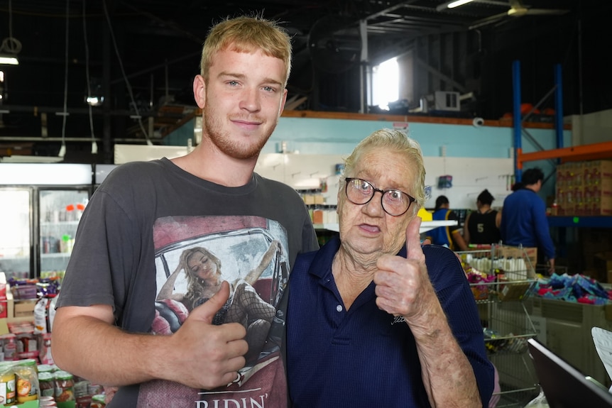 A young man and an older woman pose for a photo in a low-cost grocery store. They both wear dark t-shirts.
