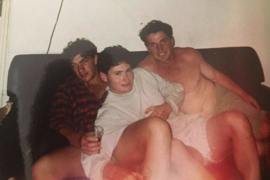 Three men in their 20s pile on a couch, one has a beer in their hand