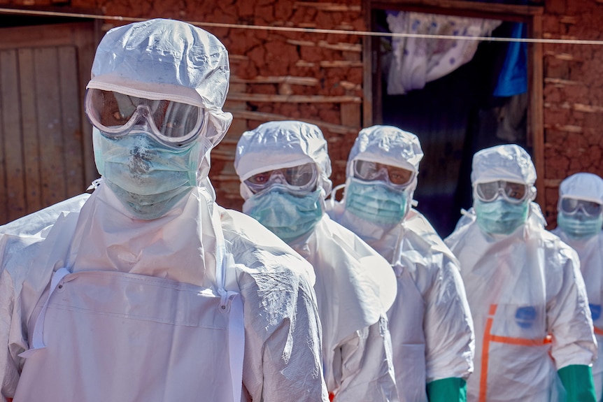 Five health care workers wearing PPE, facemasks and eyemasks standing lined up out the front of a home.