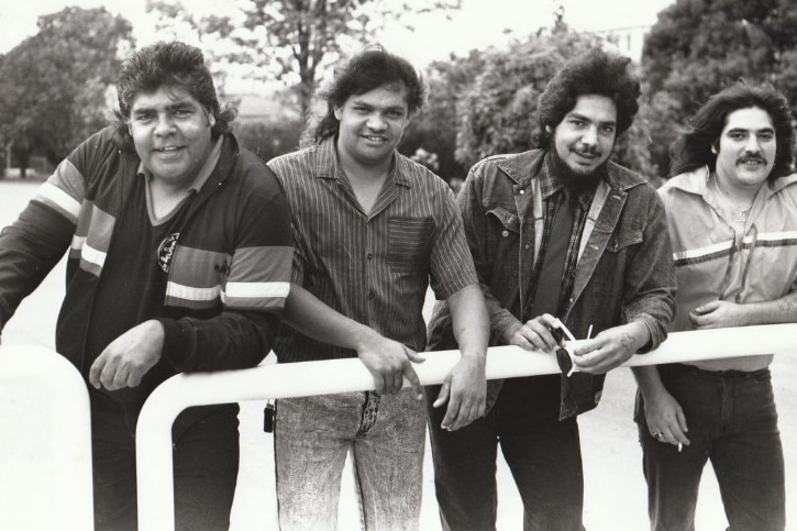 Black and white photo of four men leaning on a metal pole.