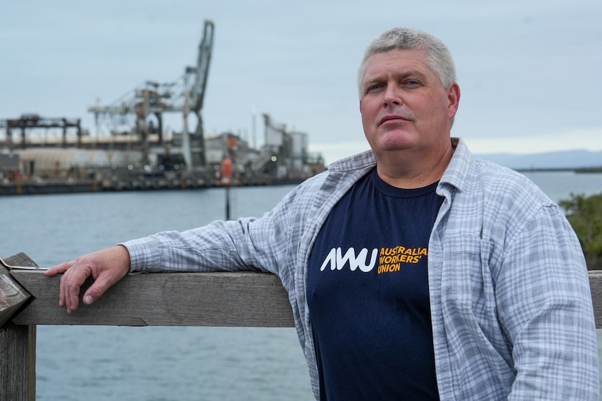 A man in an Australian Workers' Union shirt stands at a port with a metal smelter facility in the background.