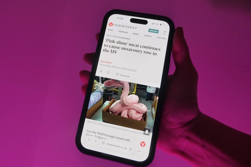 A hand holding a phone with a news article and a photo of pink slime on the screen against a pink background