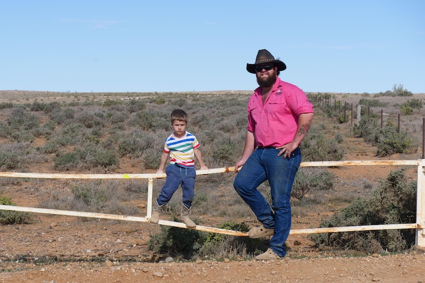 Infant boy sits on gate with his dad in outback arid location. Dad is wearing pink shirt and jeans.