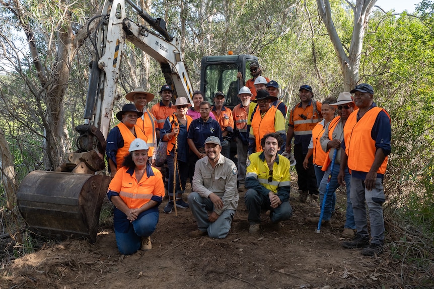 A group of people wearing high-vis vests stand in front of an excavator in the forest.